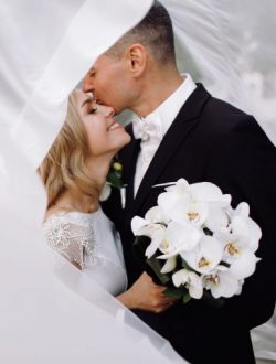 Groom in black tuxedo hugs tender stunning bride while they stand on the street of old European town